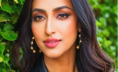 A confident woman with long dark hair and gold earrings in front of greenery.