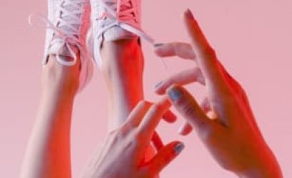 Hands reaching towards feet wearing white sneakers against a pink background.