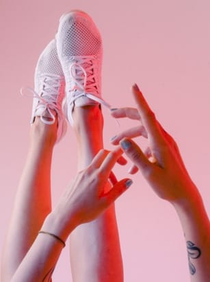 Hands reaching towards feet wearing white sneakers against a pink background.