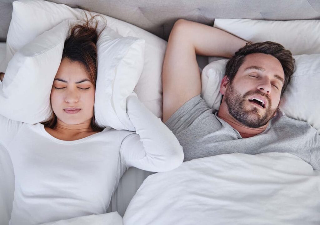 A man snoring loudly while a woman covers her ears in bed.