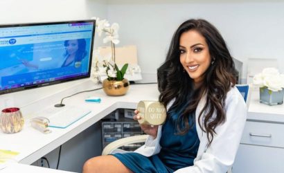 A smiling woman holding a drink at a desk with a computer and plants.