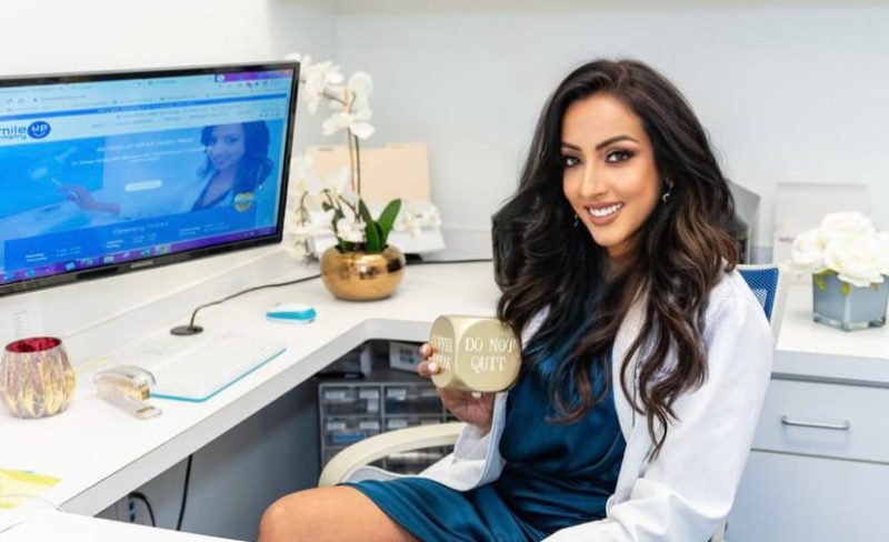 A smiling woman holding a drink at a desk with a computer and plants.