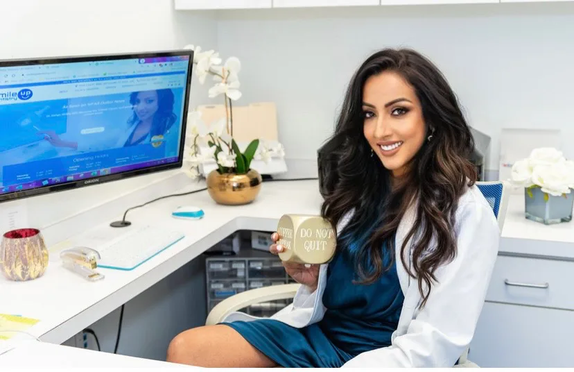 A smiling woman holding a drink at a desk with a computer and plants.